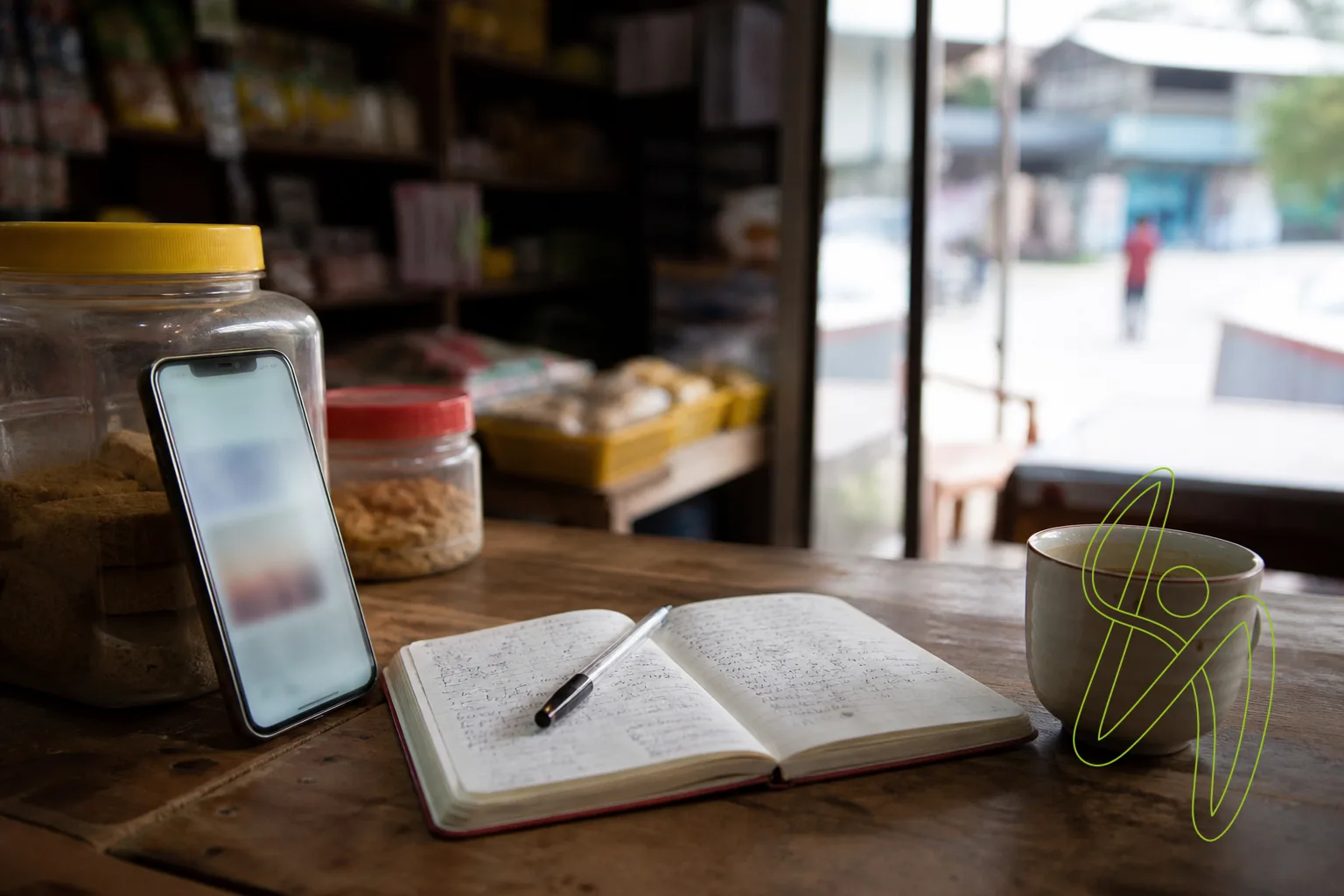 A smartphone propped on a shop counter beside a notebook and a cup of tea in morning light